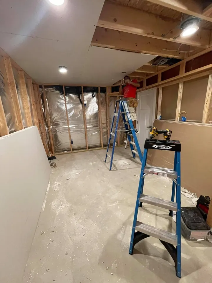 Installer on a ladder hanging ceiling drywall above exposed wall framing and insulation in a basement room.