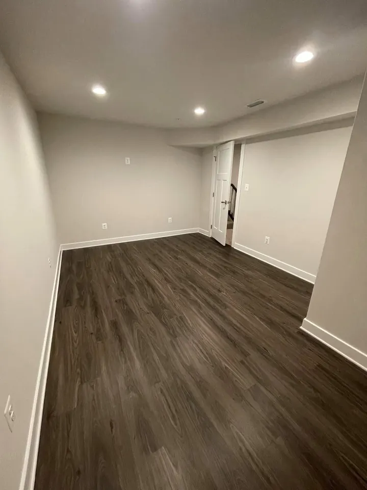 The same basement room completed with painted walls and ceiling, recessed lights, white baseboards, and dark finished flooring.